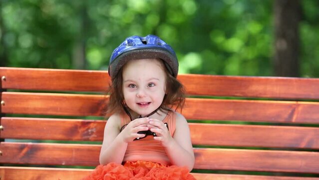 Cute little girl in blue helmet sits on bench at summer day