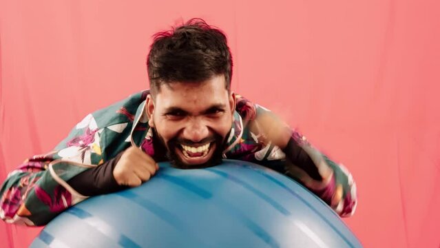 Joyful man with a playful expression laughing holding a blue exercise ball against a red background