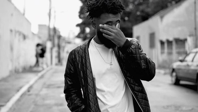 One tried young black man rubbing eyes and face with hand while walking outside in street, person struggling with chronic fatigue in monochromatic, black and white