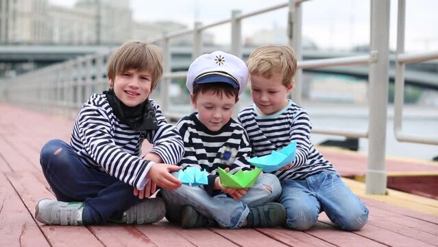 Three boys sit on the docks and playing with paper boats