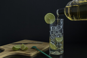 Piscola, preparing, cocktail, adding, pisco, glass with ice, wooden board, cut lemons, knife, spoon. On black table and black background. Originally from Chile, summer. Typical Chilean food and drink