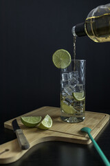 Piscola, preparing, cocktail, adding, pisco, glass with ice, wooden board, cut lemons, knife, spoon. On table and black background. from Chile, summer, vertical. Typical Chilean food and drink