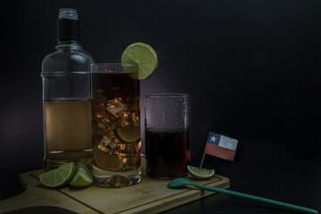 Piscola, cocktail, adding, pisco, gaseosa, glass with ice, wooden board, cut lemons, spoon, flag. On black table and black background. Originally from Chile, summer. Typical Chilean food and drink