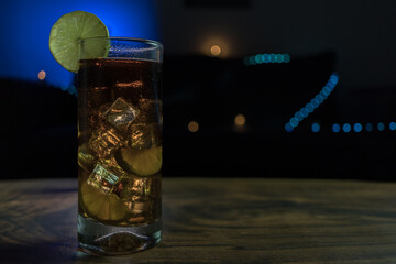 Piscola, glass, cocktail, pisco, cola, ice cubes, cut lemons. On wooden table and black background...