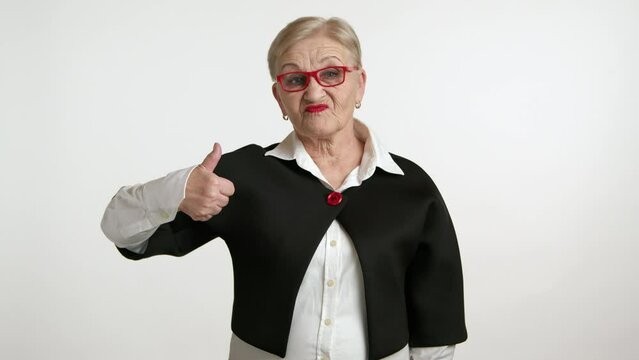 Pretty elderly woman with blonde short hair, red lipstick in red-rimmed glasses wearing white shirt and black jacket showing thumb up and thumb down. High quality 4k footage