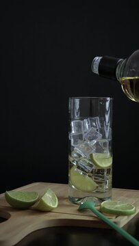 Piscola, preparing, cocktail, adding, pisco, glass with ice, wooden board, cut lemons, knife, spoon. On black table and black background. Originally from Chile, summer, vertical, video. Typical Chilea