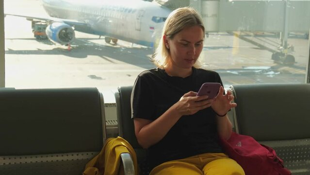 Airport terminal. Woman waiting for flight using smartphone. Girl with cell phone in airport surfing internet social media apps. Traveling female in boarding lounge of airline hub. Traveling girl