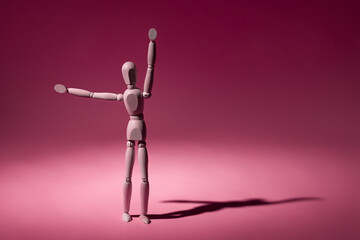A wooden mannequin with arms wide open, standing, casting a shadow on a pink background.