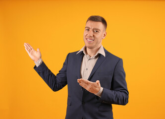 A man in a suit gesturing with one hand, smiling towards the camera on an orange background.