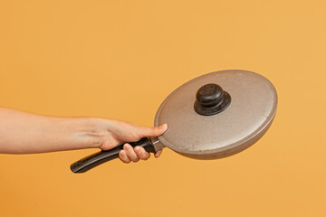 Obraz premium A hand holding a vintage-style frying pan with a black handle and lid against an orange background.