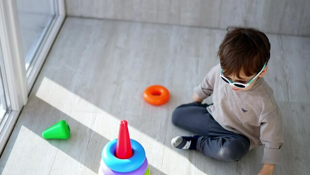 Active Caucasian toddler wearing sunglasses sits on the floor. Baby boy plays with pyramid. Top view.