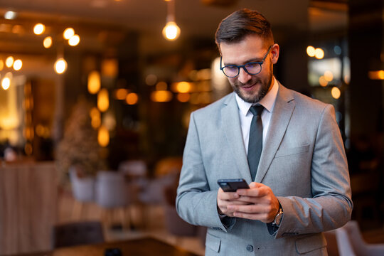Young businessman using a smartphone in a co-working office. Modern businessman sending a text message to his business clients. Well dressed businessman standing alone in a modern workplace.