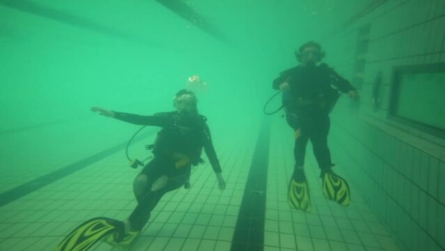 Two divers with oxygen tanks train underwater in pool 