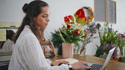 Serious florist work laptop digital device in flowers store. Focused woman work.