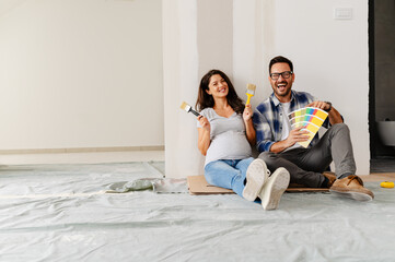 Obraz na płótnie Canvas A young, beautiful, and cheerful couple, consisting of a man and his pregnant wife, are seated on the floor of an empty house. They are in the process of selecting colors for decorating their new home