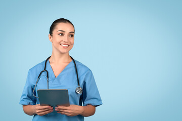 Cheerful young woman nurse in blue medical uniform holding digital tablet and looking thoughtfully...