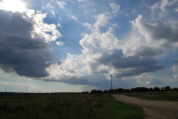 Rural sandy road. The road passes under a gloomy sky with white-gray clouds. Tall green grass grows...
