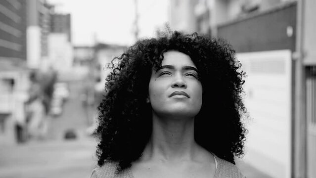 One hopeful young black woman with curly hair standing in street looking up at sky with HOPE and FAITH in black and white. African American person feeling the presence of GOD