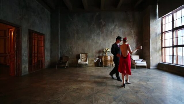 Girl in red dress and man in black suit dance tango in old room