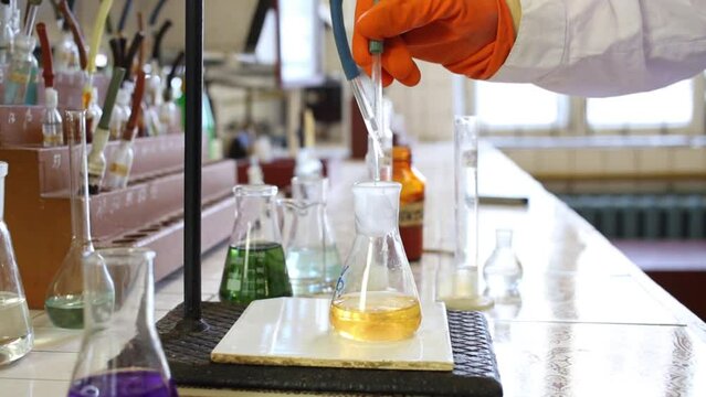 Hands in gloves drips liquid into flask with yellow liquid in lab