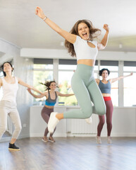 Girl engaged shake leg hurdle step in fitness studio with female friends, group of youthful people jumped into air. Young women in good mood actively move and dance at choreography lesson.