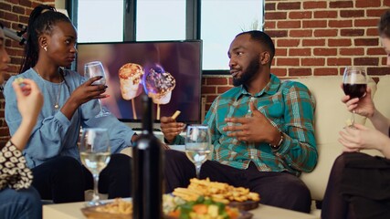 Multicultural group friends celebrating african american man birthday, drinking wine and eating snacks in apartment. Guests laughing and talking at house gathering after offering host presents