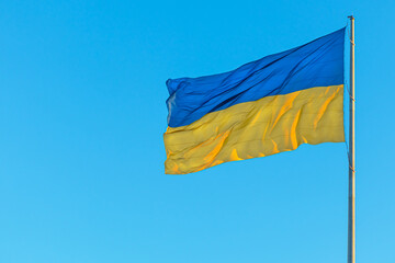 The large national flag of Ukraine flutters in the wind. Yellow-blue flag on a flagpole against the background of a clear blue sky on a sunny day