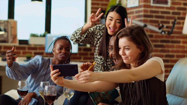Asian, african american and caucasian people taking group pictures with mobile phone at apartment party. Friends in stylish home smiling for selfie photographs with wine and champagne glasses in hand