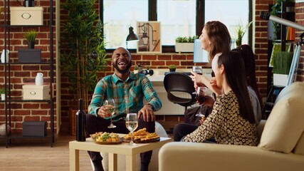 Best friends laughing together, enjoying alcohol beverages and eating from charcuterie board in cozy apartment living room. BIPOC group of people talking at home, entertaining each other