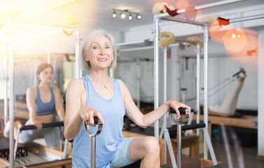 Fototapeta premium Portrait of positive focused aged woman doing remedial gymnastic in pilates studio, exercising on wunda chair..