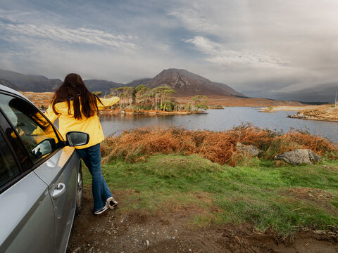 Teenager girl in yellow tourist jacket standing by silver car. Twelve pine island and stunning mountains in the background. Connemara County Galway, Ireland. Travel and sightseeing, Irish landscape