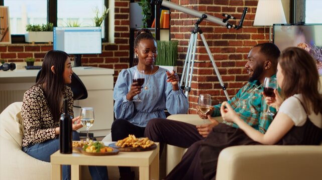 Multiracial friends at apartment party paying attention to interesting story shared by african american woman. BIPOC group of people listening to guest while enjoying drinks and charcuterie platter