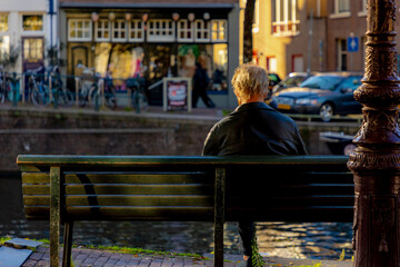 Obraz premium Backside of a old man on wooden bench with blurred view along the canal, The senior guy sitting and relaxing, Hobby and leisure activity of retirement age, Amsterdam capital city of the Netherlands.
