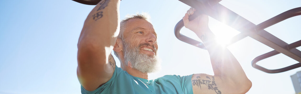 Strong and healthy people. Happy bearded senior man pulling up on horizontal bar and smiling while training outdoors. He is working out at the stadium. Morning exercises