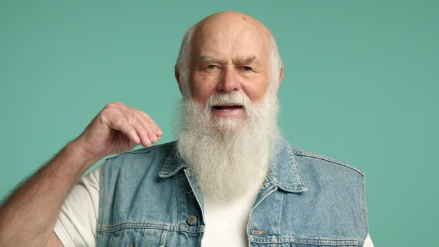 A mature gentleman wearing a denim vest humorously dismisses idle chatter with a single hand motion, set against a serene turquoise background. Camera 8K RAW. 