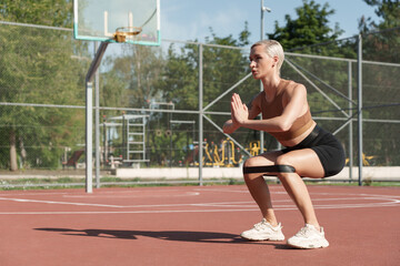 An athletic woman engages in a powerful squat workout with resistance bands at a sunlit basketball...