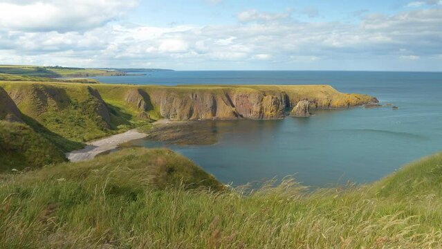 Sheltered sandy beach nestled between high rocky cliffs overgrown with grass. Clifftop grass gently rustling in summer breeze as clouds roll in over the picturesque coastline of eastern Scotland.