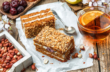 Two slices of delicious honey biscuit cake with cream, apples, cherries and peanuts on wooden background with cup of tea and spring flowers. Sweets, dessert and pastry, homemade cakes, top view
