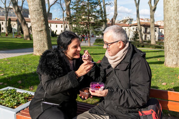 Fototapeta premium Mature romantic couple sitting on bench in park and enjoying a sweet and delicious cake. Street foods are popular among tourists and people. Food concept.