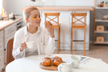 Young woman with cup of coffee eating bun in kitchen