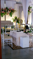 Place for wedding ceremony in white color ,with white fireplace and chandeliers decorated with flowers and white cloth and wooden chairs for guests on each side outdoors.