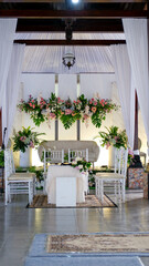 Place for wedding ceremony in white color ,with white fireplace and chandeliers decorated with flowers and white cloth and wooden chairs for guests on each side outdoors.
