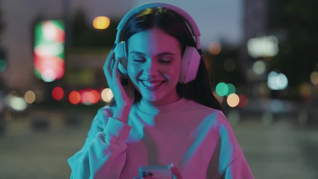 A joyful young woman in a modern setting, headbanging to music in her headphones with vibrant neon city lights at night as a backdrop.