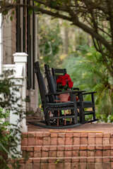 A porch or stoop with two wooden rockers, a small table, and a pot of colorful red flowers. Brick steps and white rail lead to the patio. The garden has large lush green plants, trees, and shrubs. 