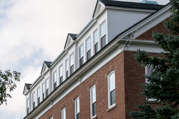Fototapeta premium The exterior of a large multi-level university brown brick building with rows of multiple glass windows. The shingled white wooden roof has a row of peaked dormers with double-hung glass windows. 