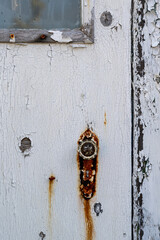 A white wooden door with a vintage decorative glass door knob. The exterior of the antique house is white clapboard walls. There's rust staining the door and the old metal faceplate of the handle.