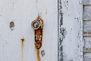 A white wooden door with a vintage decorative glass door knob. The exterior of the antique house is white clapboard walls. There's rust staining the door and the old metal faceplate of the handle.