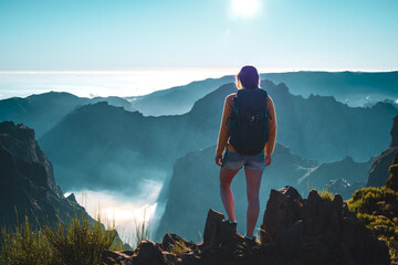 Backpacker tourist stands on the edge of a deep, cloud-covered valley and enjoys the breathtaking panoramic view of the volcanic mountain landscape. Pico do Arieiro, Madeira Island, Portugal, Europe. - obrazy, fototapety, plakaty