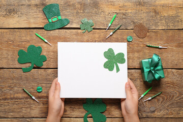 Female hands with blank card, gift box and decor on wooden background. St. Patrick's Day celebration