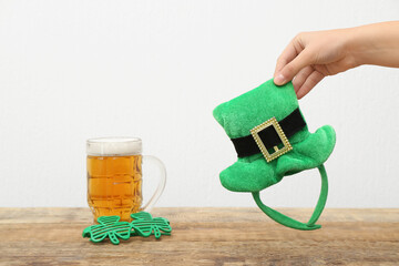 Female hand with leprechaun hat, mug of beer and plastic eyeglasses on wooden table against white...